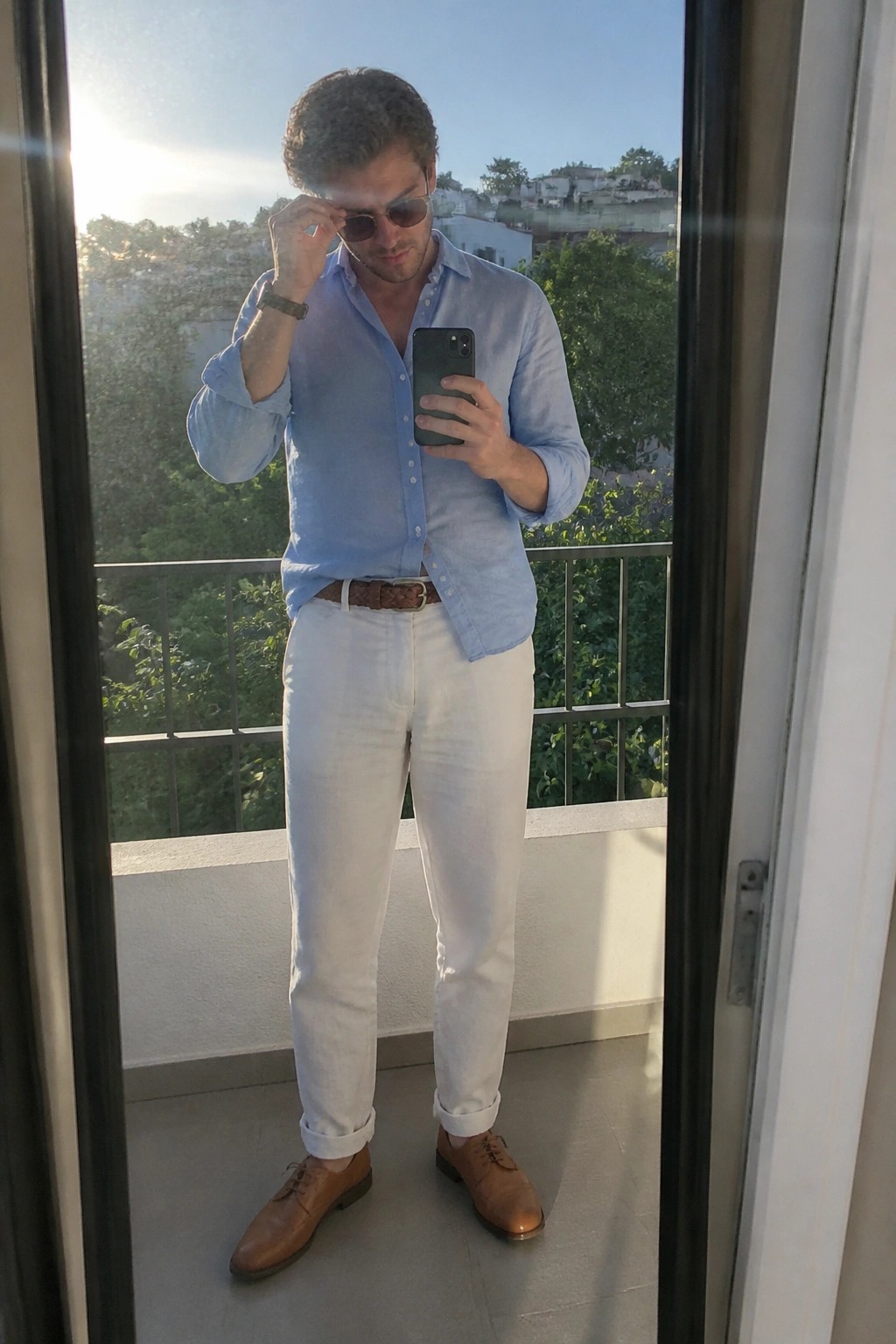 Man wearing a light blue linen button-up shirt tucked into white pants with a brown woven belt, brown leather dress shoes, sunglasses, and a watch while holding a phone.