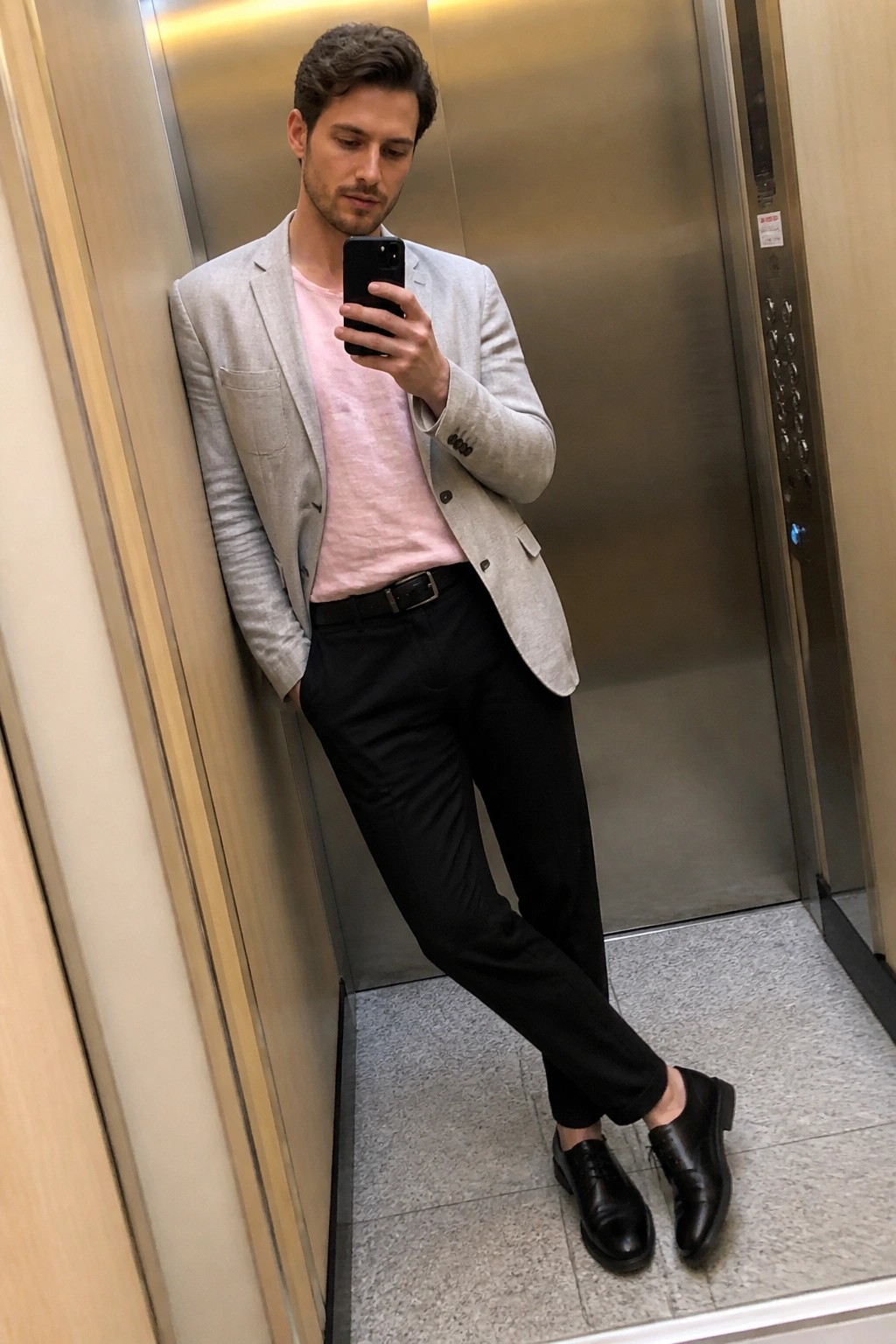 Man in light gray blazer over pink tee taking elevator mirror selfie