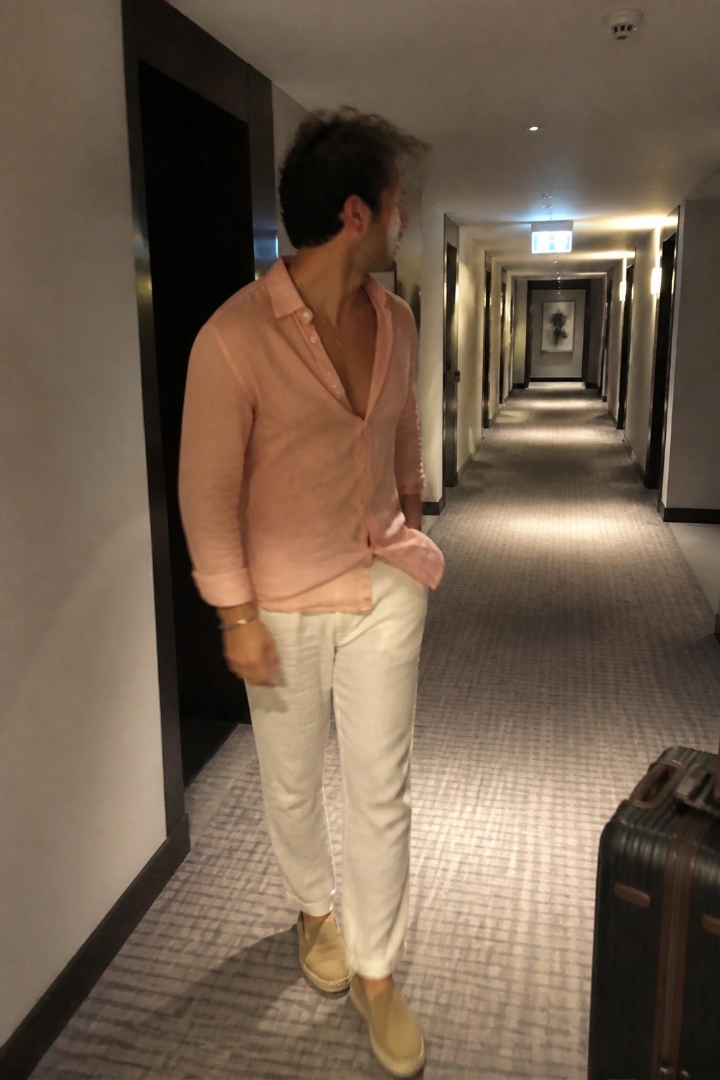 Man in a light pink linen button-up shirt with white pants and beige slip-on shoes standing in a hotel hallway next to a dark suitcase.