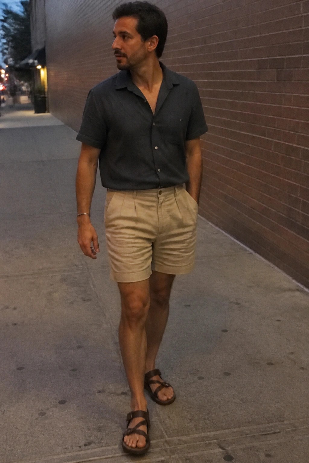Man in dark gray short sleeve button up shirt, beige pleated shorts, and brown strappy sandals standing on a sidewalk.