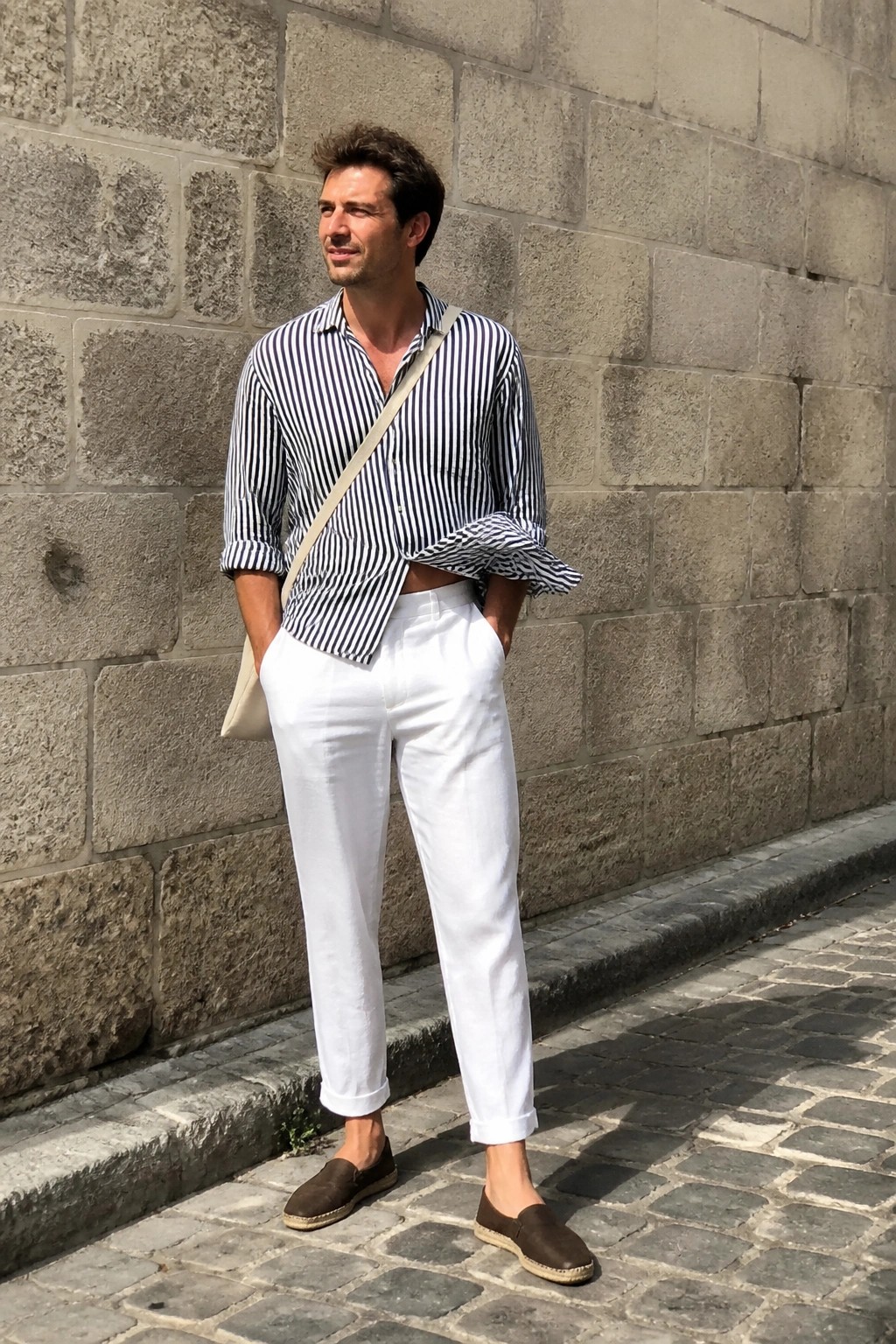 Man in striped shirt and white pants against stone wall on cobblestones.