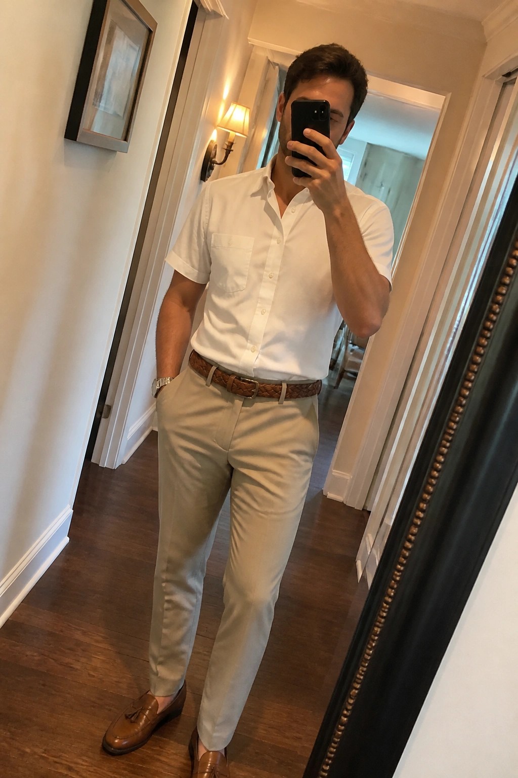 Man wearing white short sleeve button down shirt tucked into light khaki tailored pants with brown woven belt and brown leather tassel loafers while holding a phone for a mirror selfie.