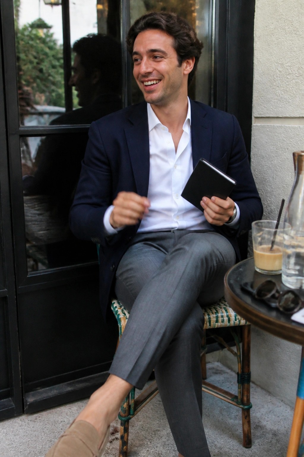 Man in navy blazer and white shirt smiles while holding notebook at cafe table.