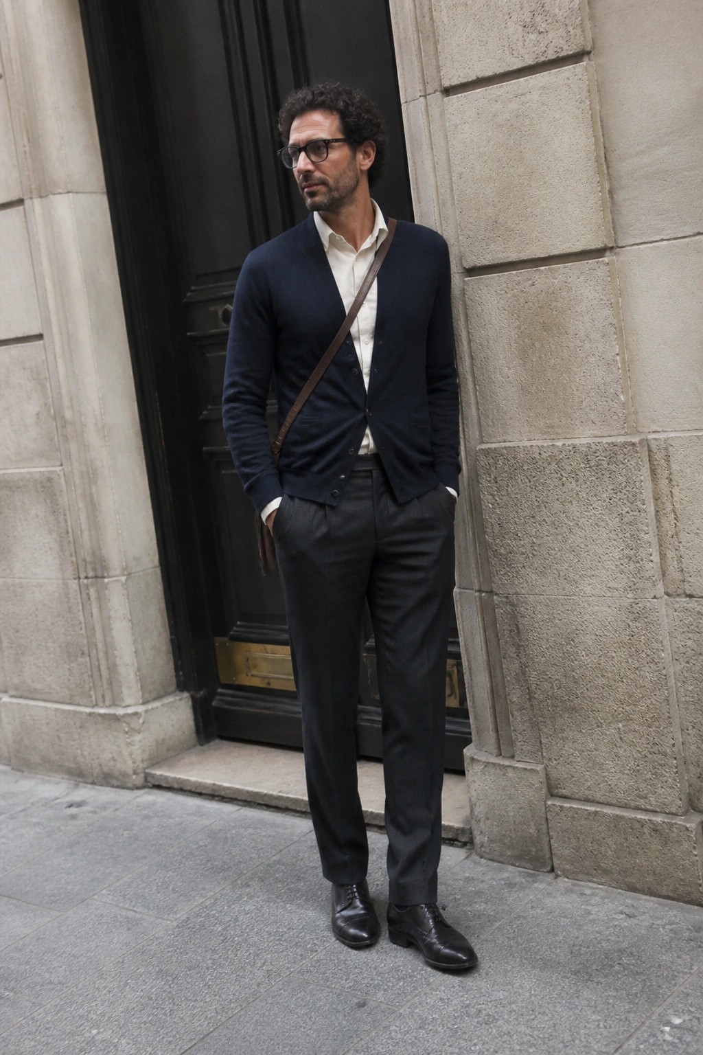 Man in navy cardigan and gray trousers leaning against stone wall, hands in pockets.