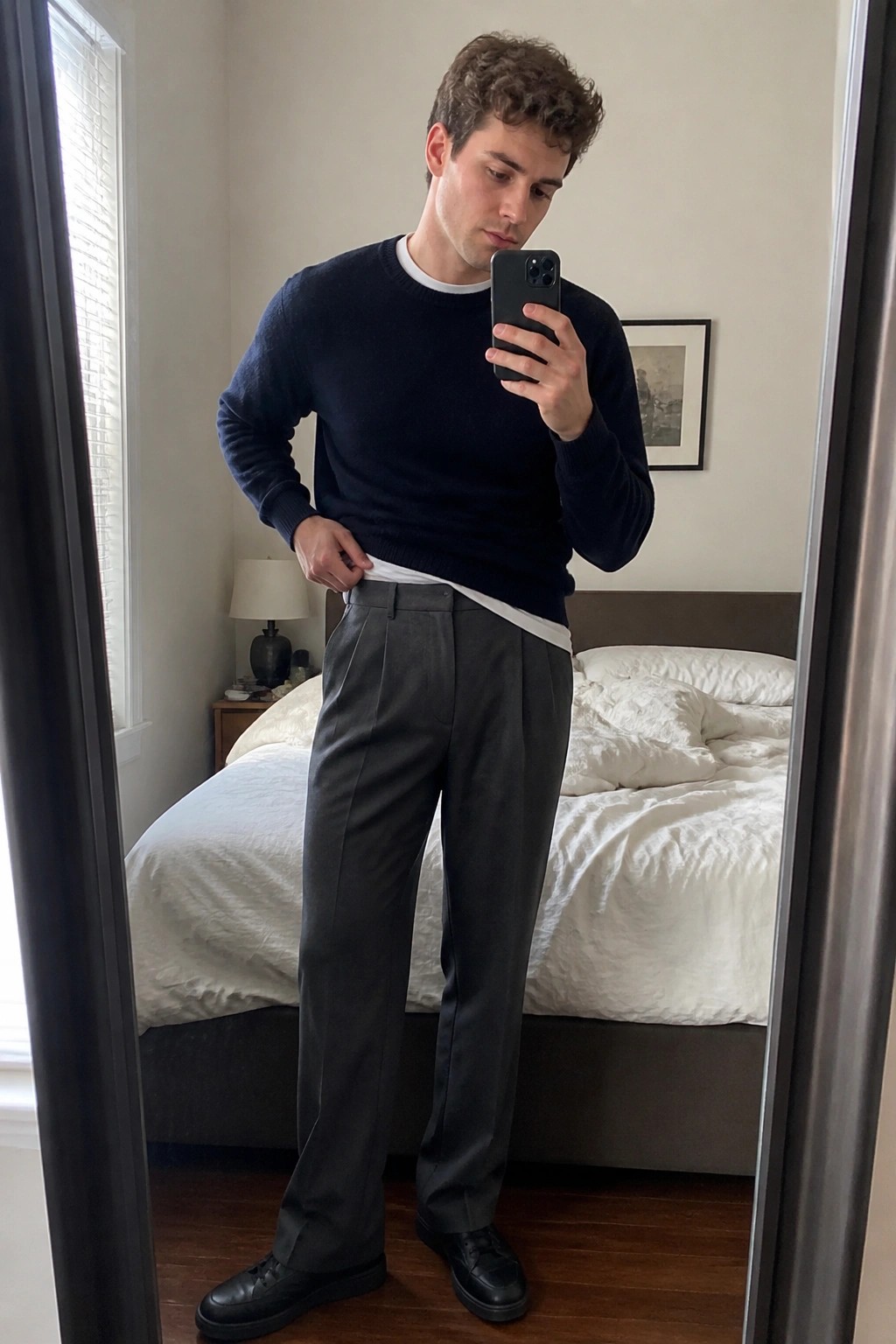 Man in navy sweater and gray pleated trousers taking bedroom mirror selfie