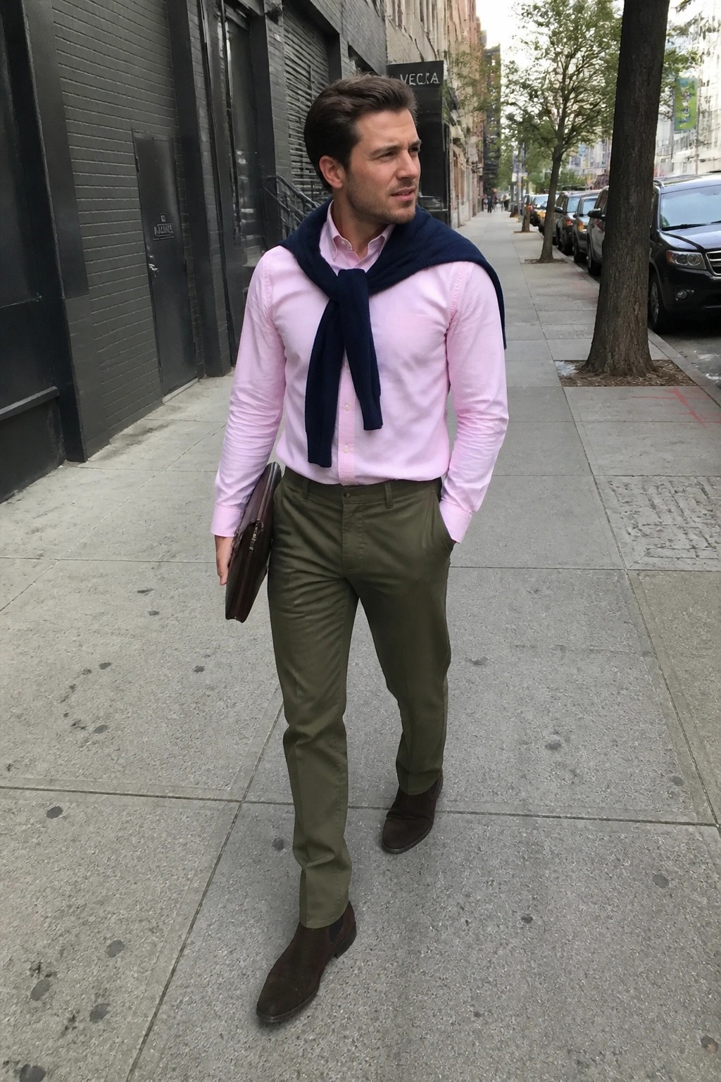 Man in pink shirt and green pants with navy sweater on city sidewalk