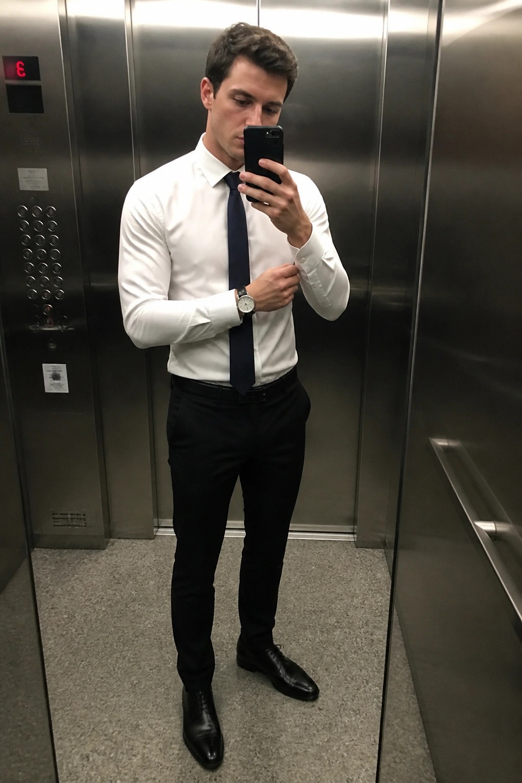 Man in white shirt and navy tie taking mirror selfie in elevator.
