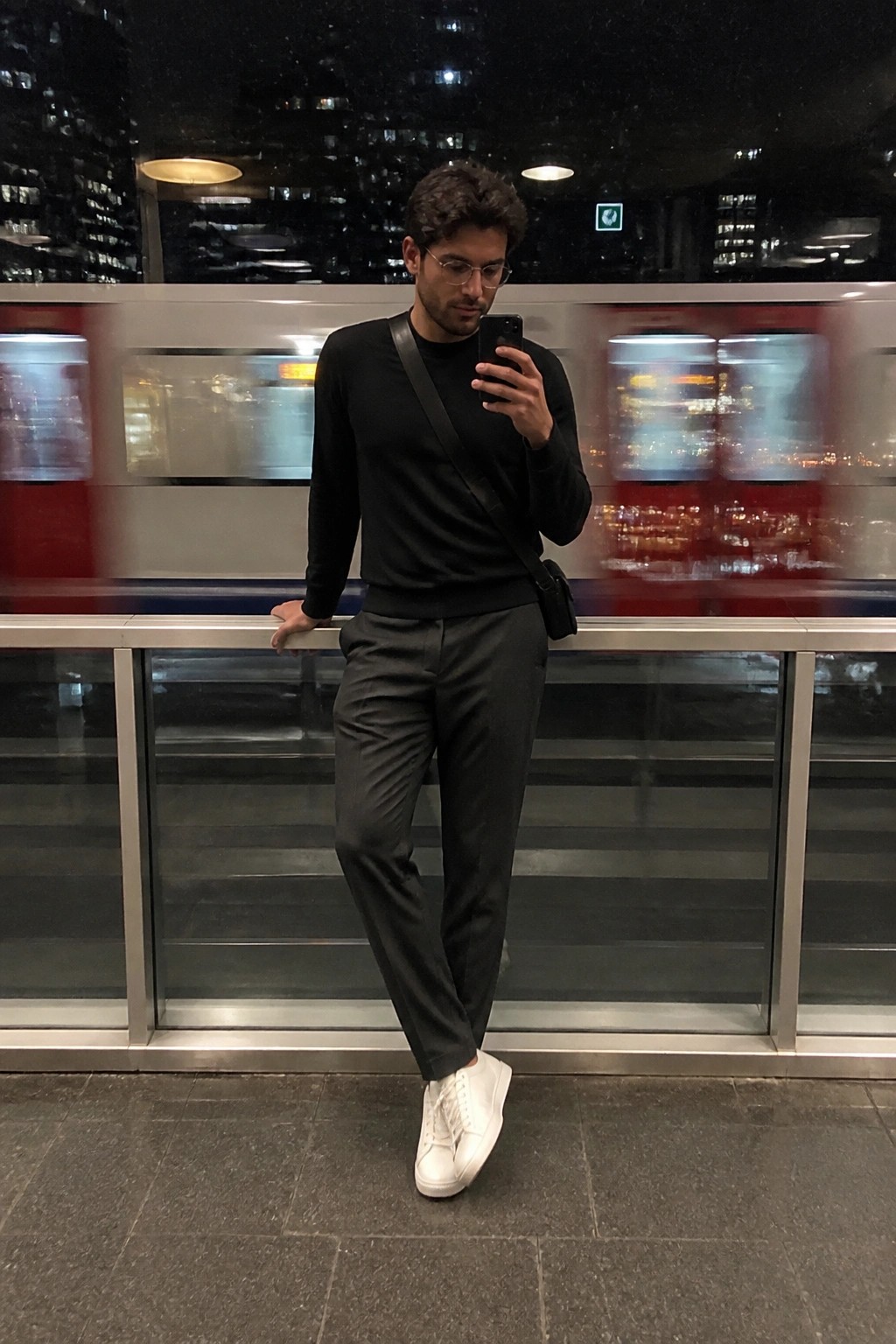 Man in black sweater and gray pants checks phone by blurred subway train at night