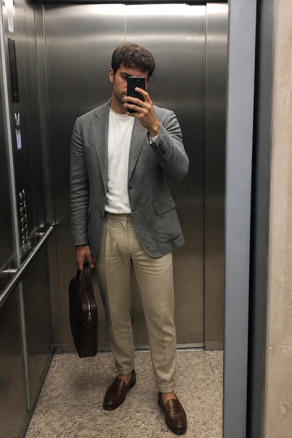 Man in gray blazer, white tee, beige pants, and brown loafers holding phone and bag in elevator