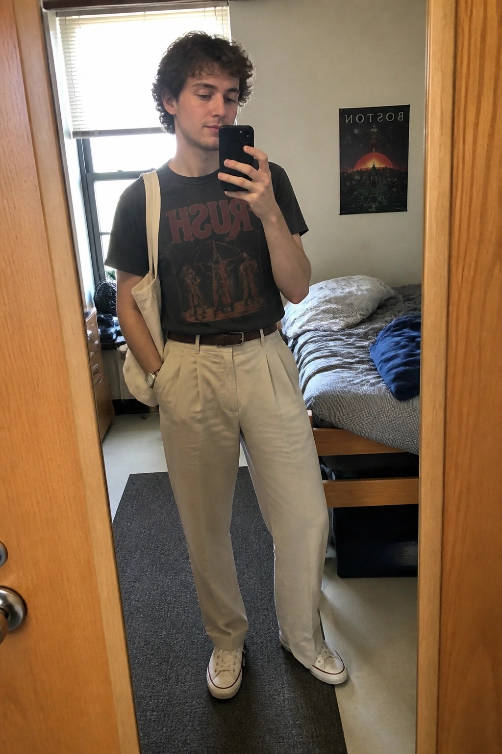 Young man in gray Rush t-shirt and beige pants takes mirror selfie in dorm room.