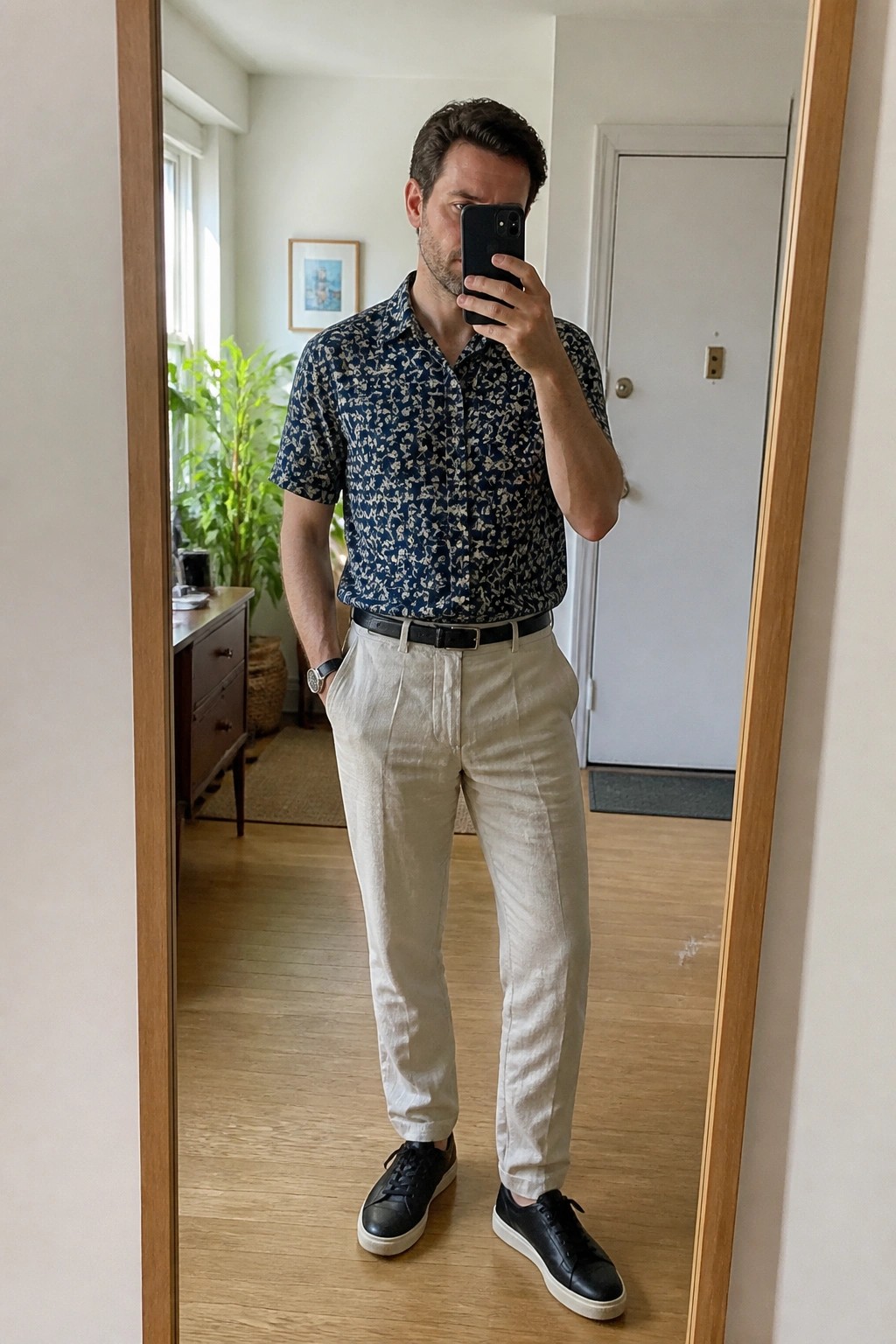 Man in patterned shirt and beige pants taking mirror selfie indoors.