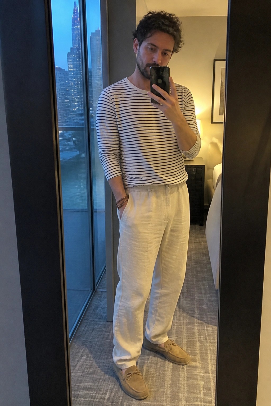 Man in white linen pants, navy striped long sleeve shirt, and beige suede loafers taking a mirror selfie indoors.