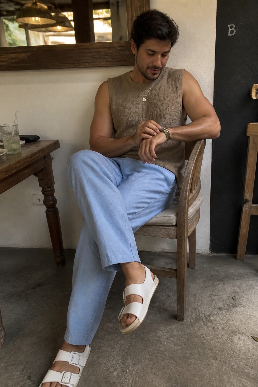 A man in light blue linen pants, a beige sleeveless knit top, white double-strap sandals, a watch, and a necklace seated on a wooden chair.