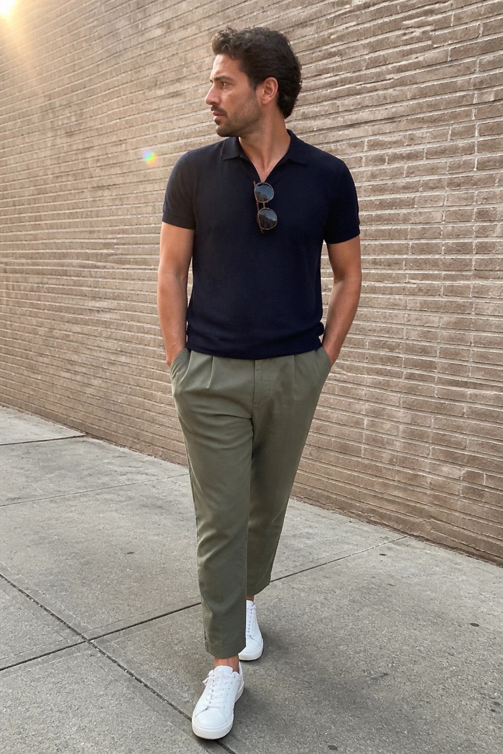 A man wearing a navy polo shirt tucked into olive green pants with white sneakers and sunglasses hanging from the collar, standing against a brick wall.