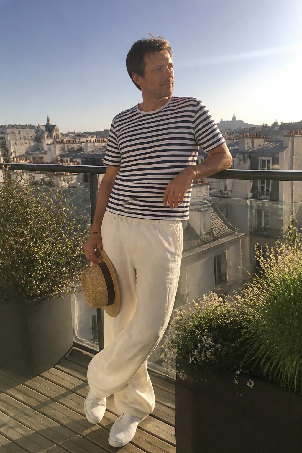 Man wearing navy and white striped short sleeve t-shirt tucked into loose white linen pants with white sneakers and holding a straw hat while standing on a wooden deck.