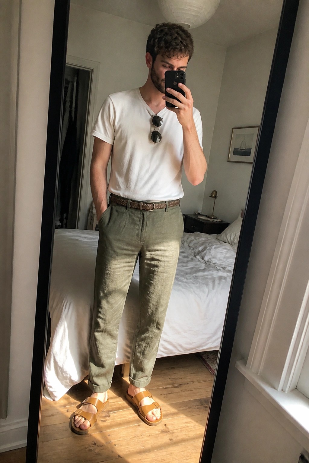 Man in white t-shirt and green pants taking mirror selfie in bedroom