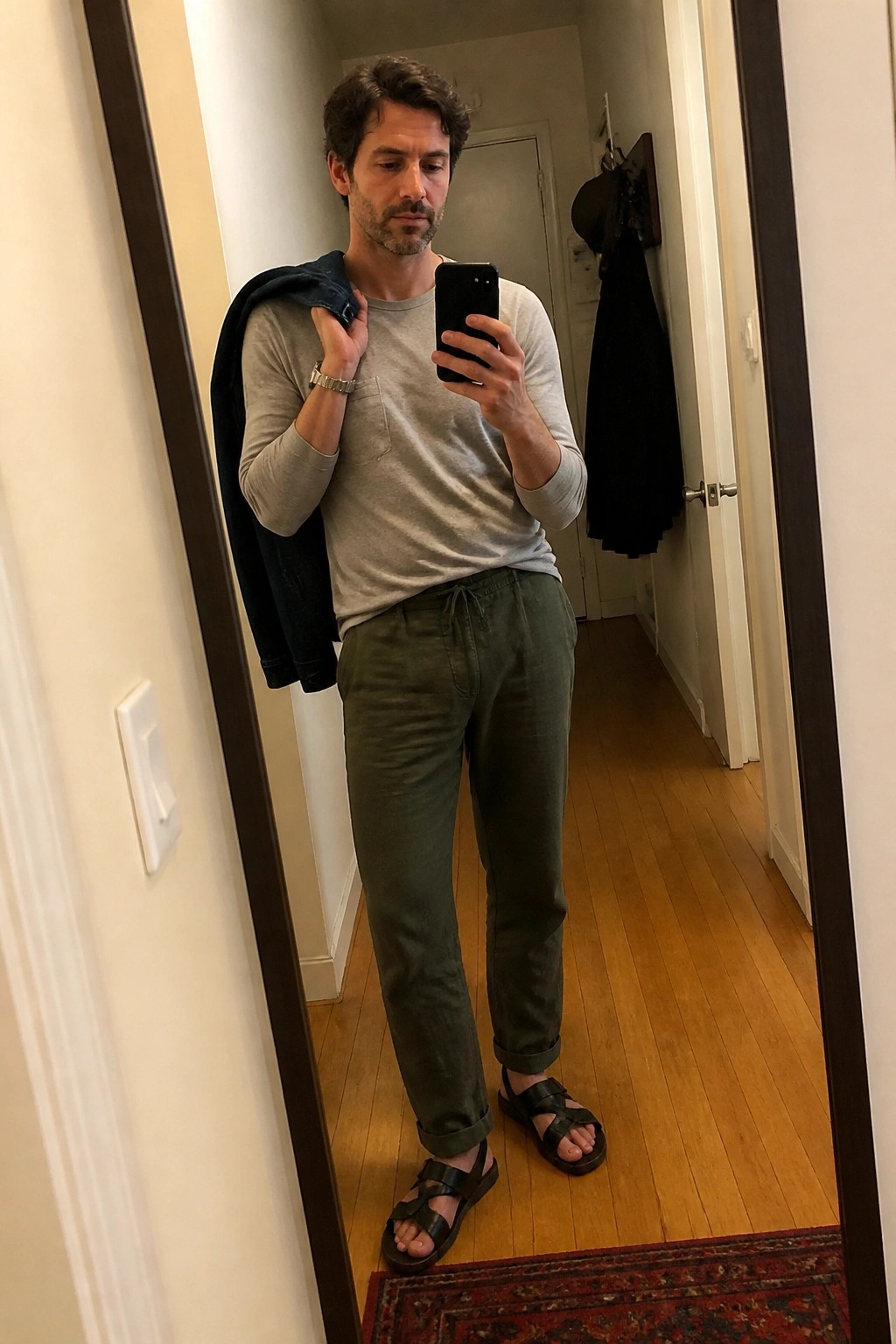 Man in gray shirt and green pants holds jacket for mirror selfie in hallway