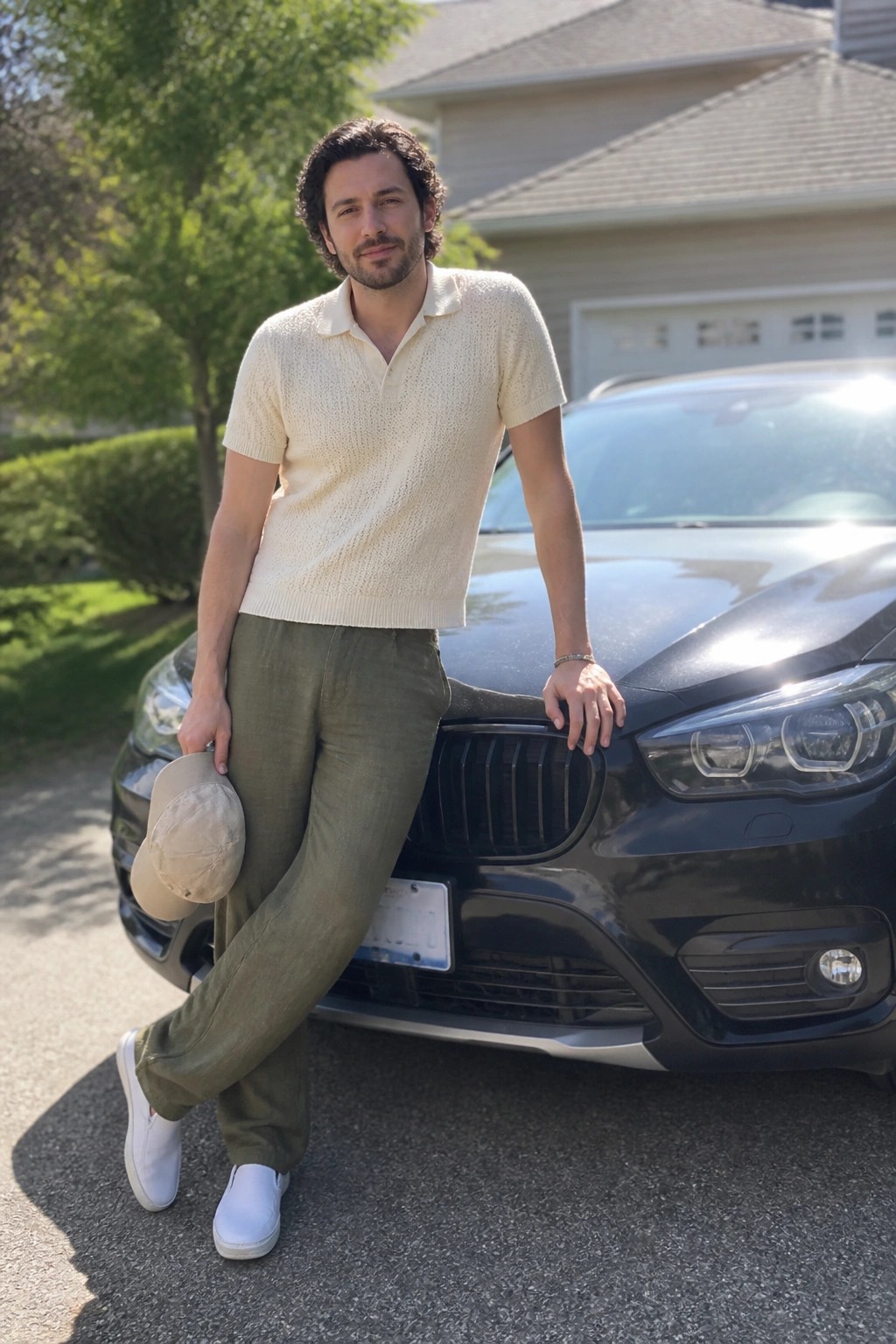 Man in cream knit shirt and green pants leaning on black BMW car