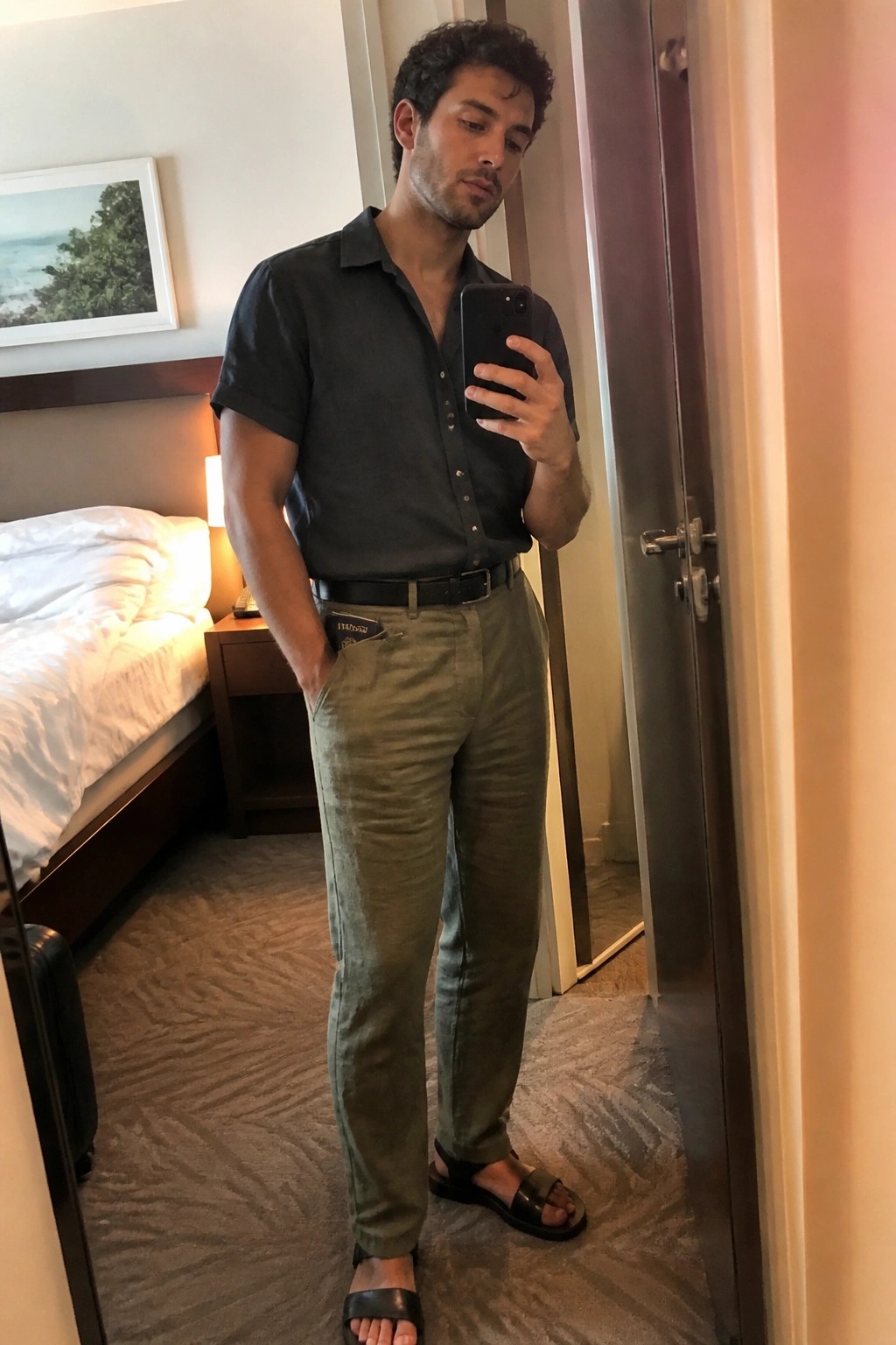 Man in dark shirt and olive pants taking mirror selfie in hotel room