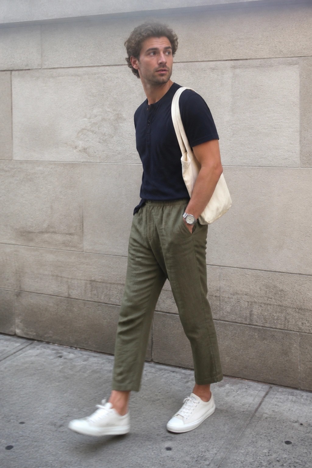 Curly-haired man in navy henley and olive pants with white tote bag on street.