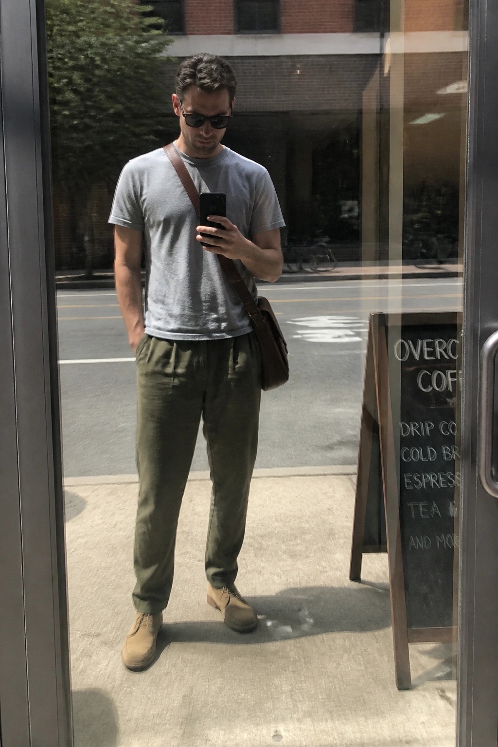 Man in gray t-shirt and green pants checks phone in glass doorway with brown bag
