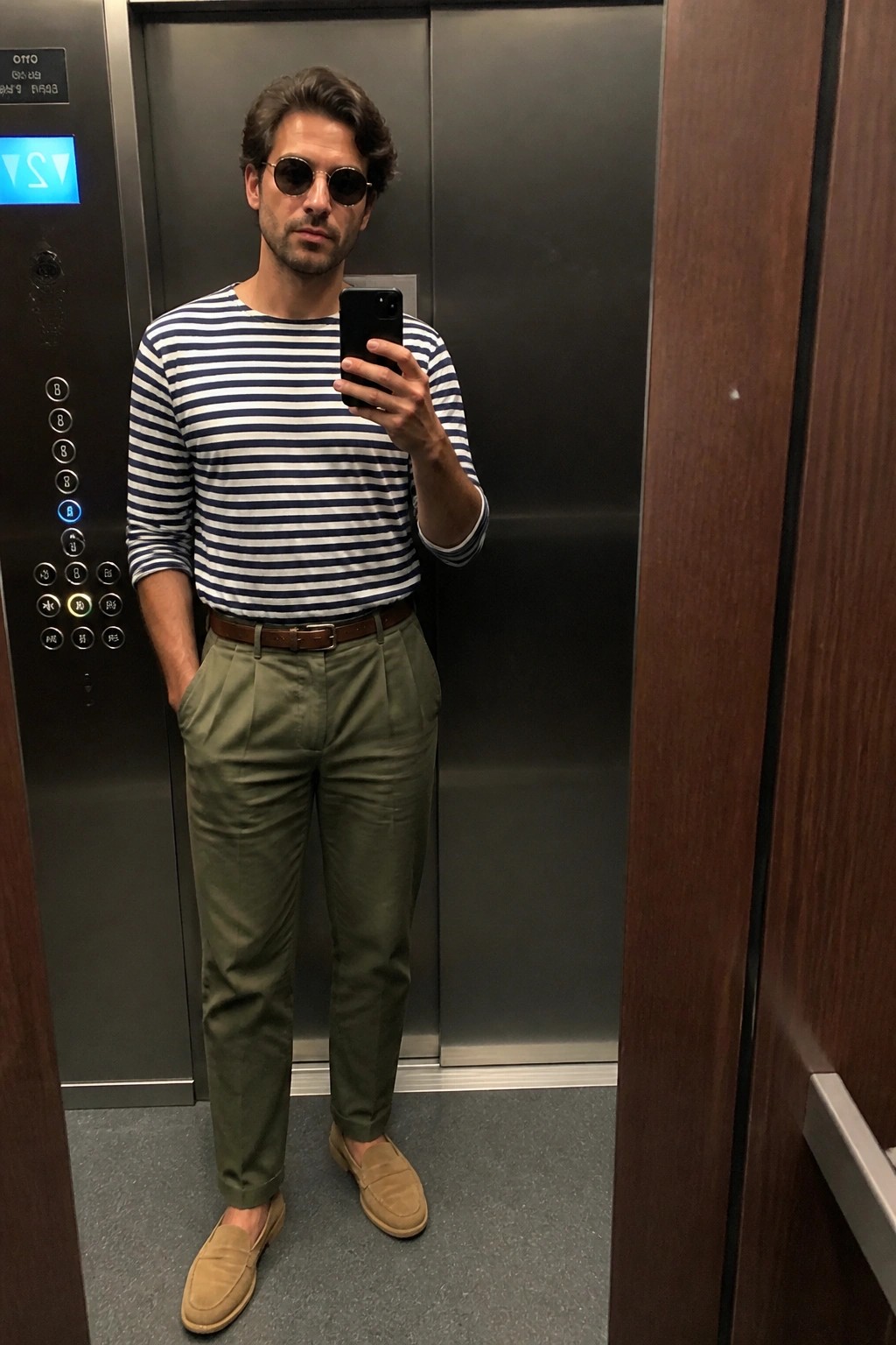 Man in striped shirt and green pants taking elevator mirror selfie.