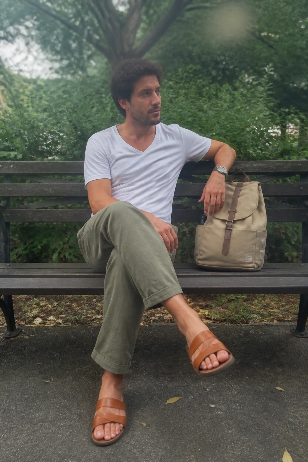 Curly-haired man in white tee and olive pants on bench beside beige backpack.