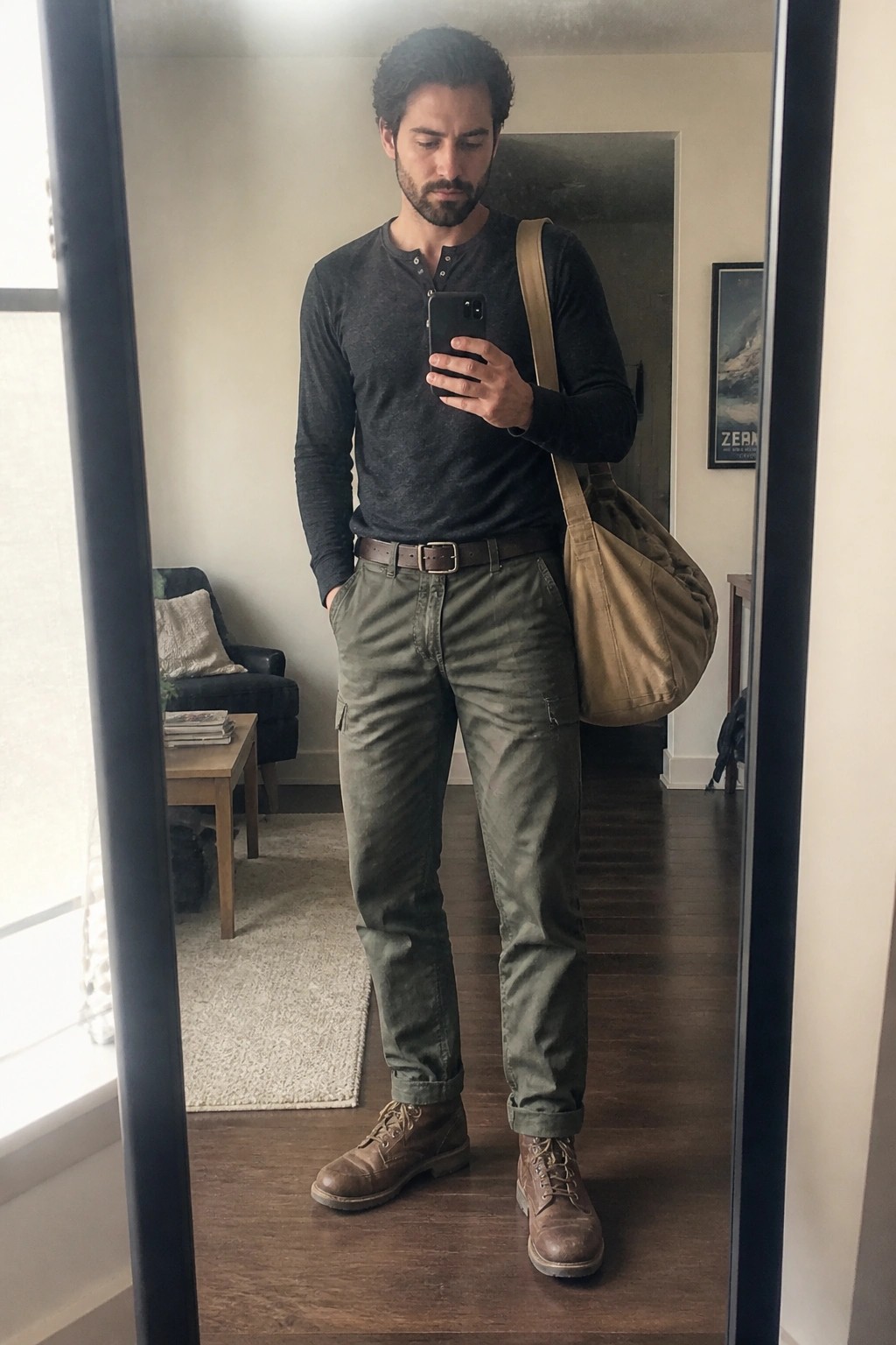 Man wearing olive cargo pants, dark gray henley shirt, brown leather boots, brown belt, and tan messenger bag while taking a mirror selfie.