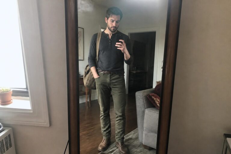 Man wearing olive cargo pants, dark gray henley shirt, brown leather boots, brown belt, and tan messenger bag while taking a mirror selfie.