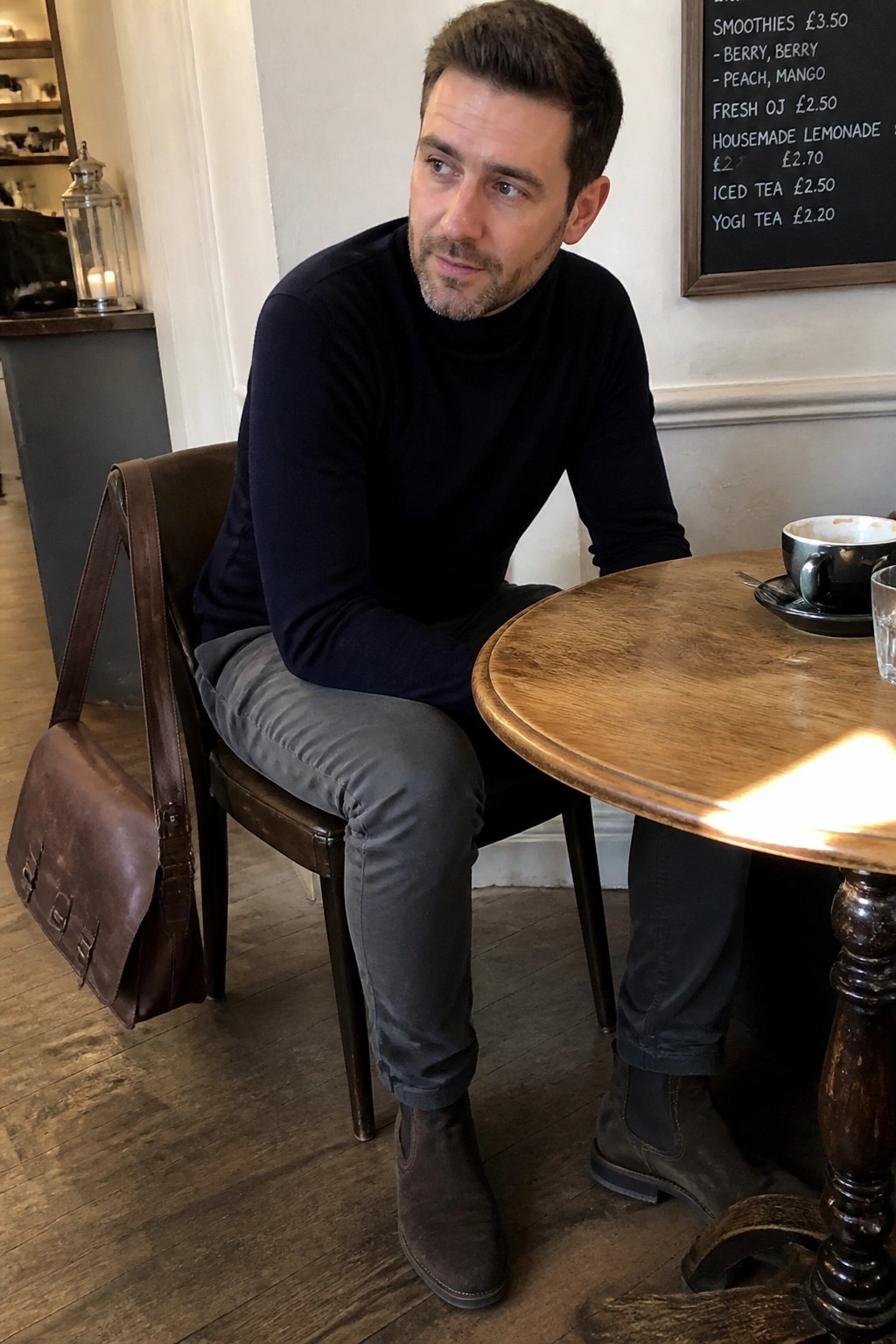 Man in navy turtleneck with leather bag at wooden cafe table