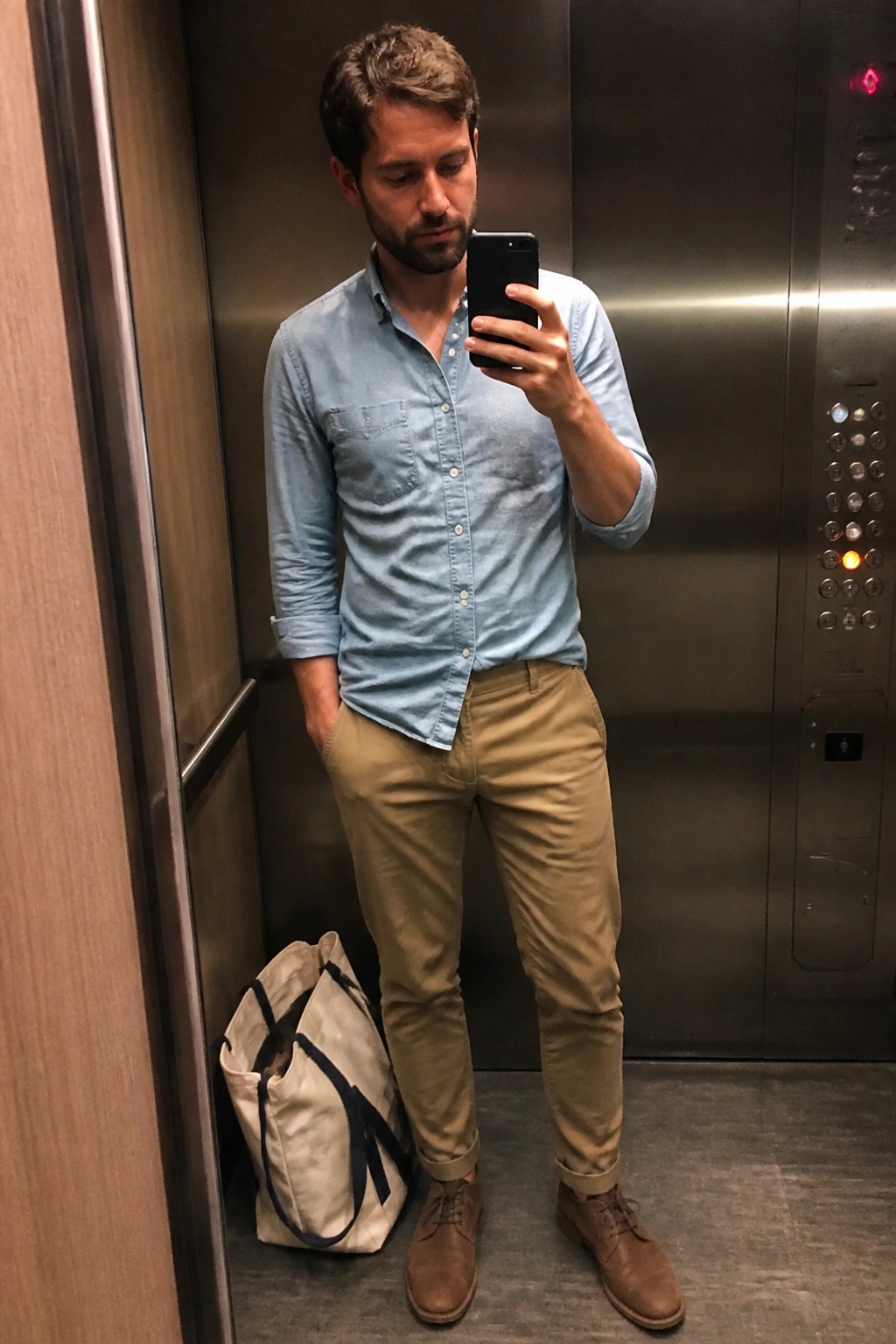 Man in blue shirt and khaki pants taking mirror selfie in elevator with tote bag.