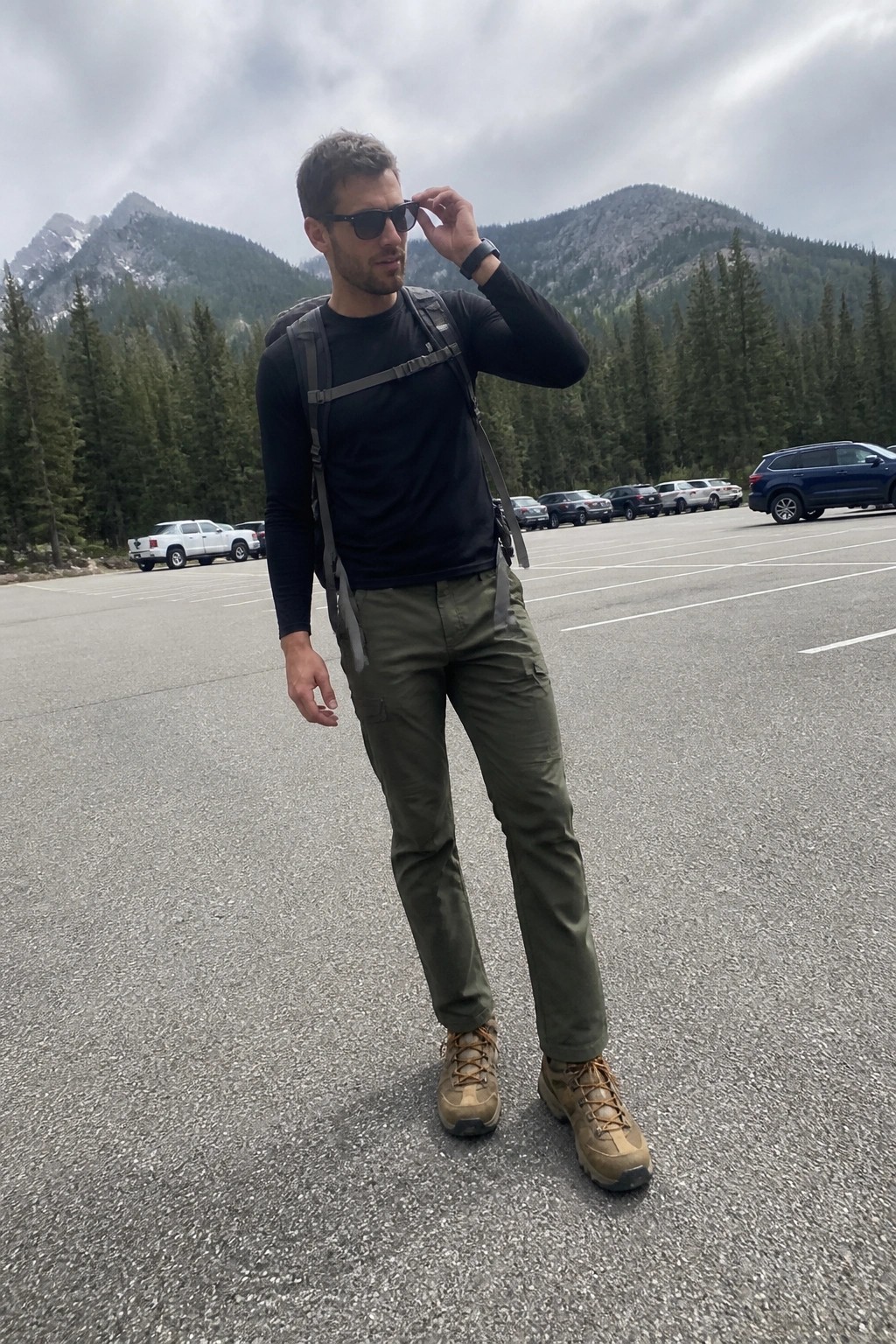 Man in olive cargo pants, black long-sleeve shirt, brown hiking boots, gray backpack with chest strap, and sunglasses standing on asphalt.