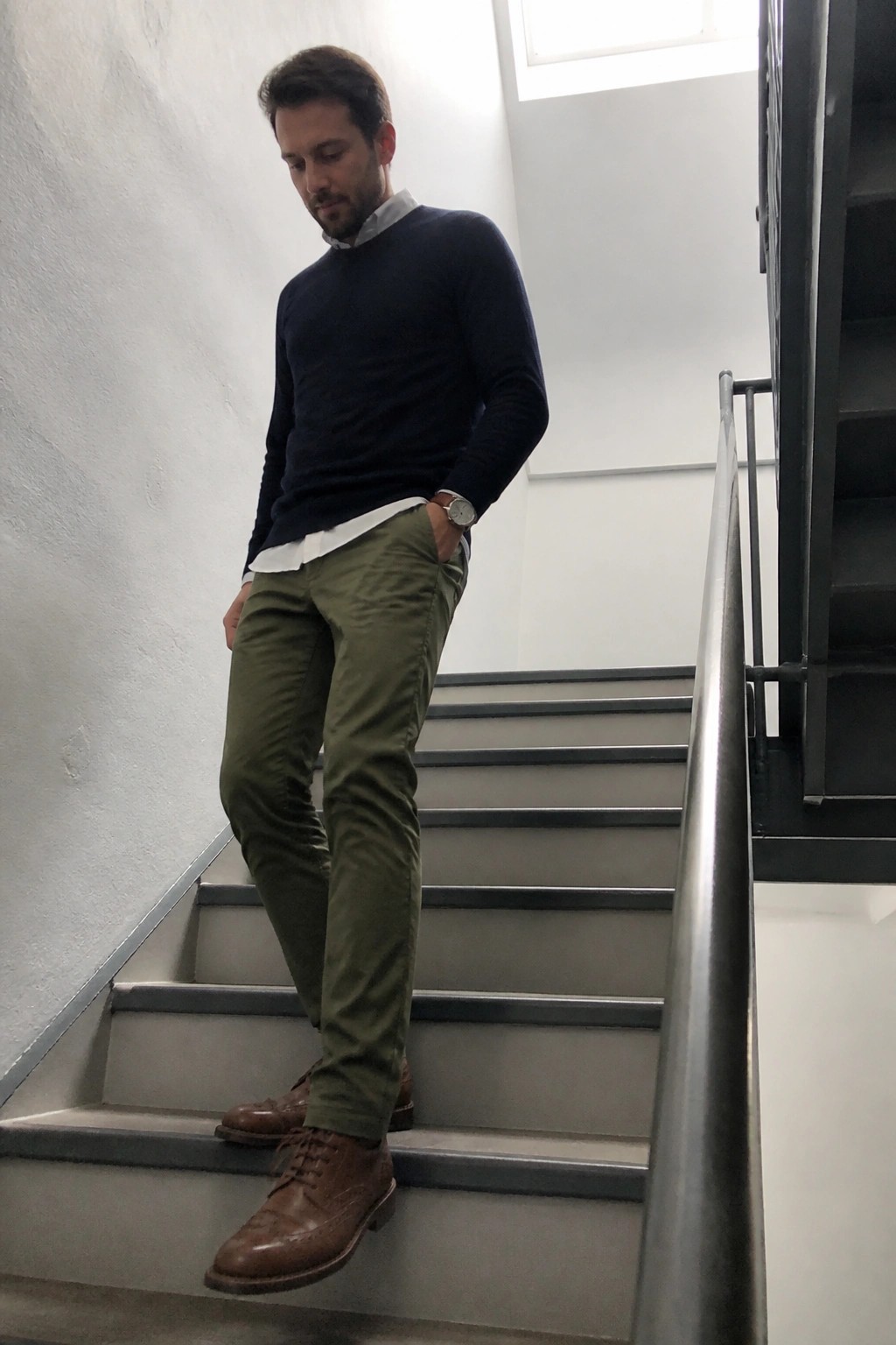 Man in navy sweater and olive pants standing on indoor staircase with hand in pocket.