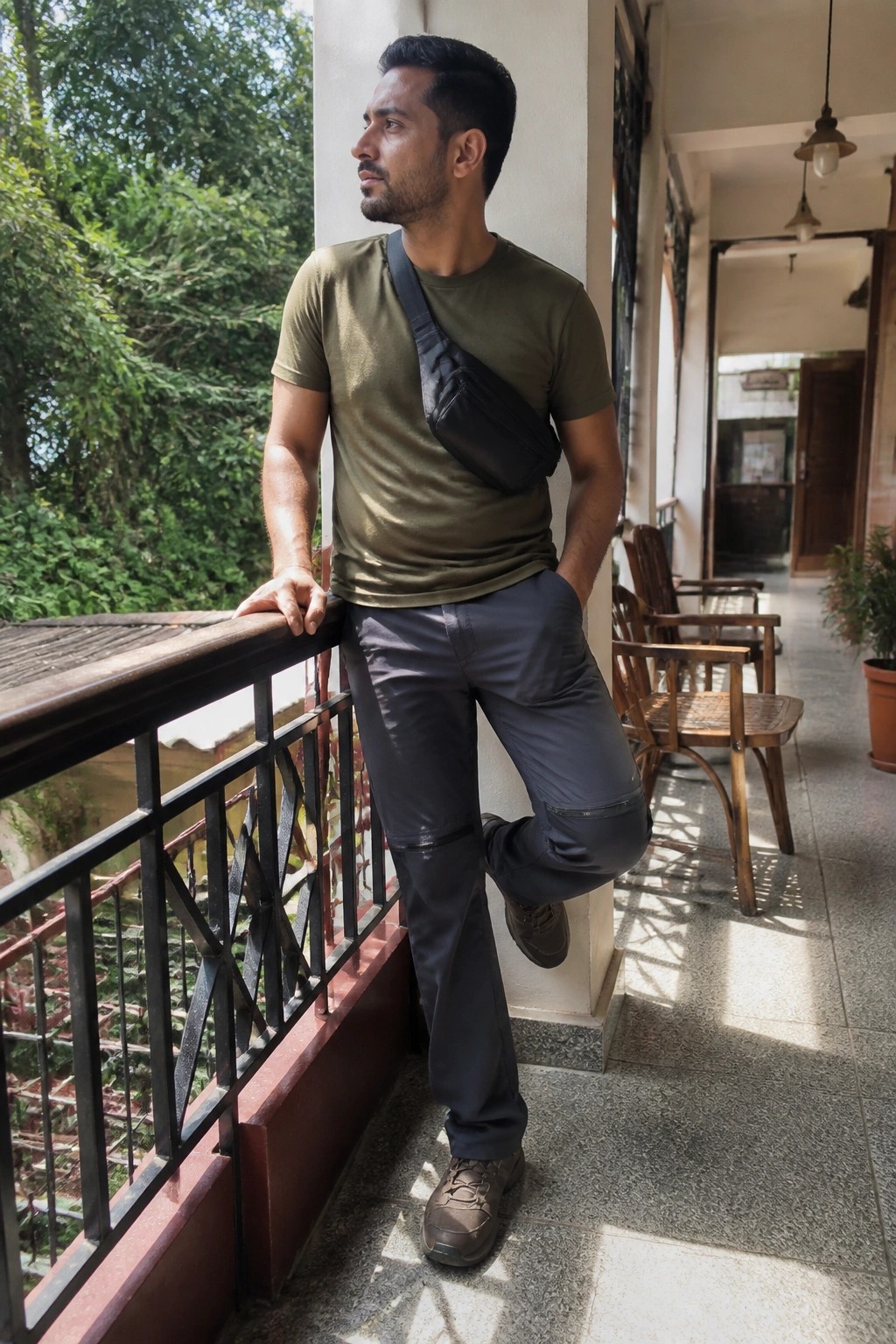 A man in an olive green short sleeve t-shirt, dark gray reinforced travel pants, brown lace up boots and a black crossbody sling bag standing casually with one foot raised on a balcony railing.