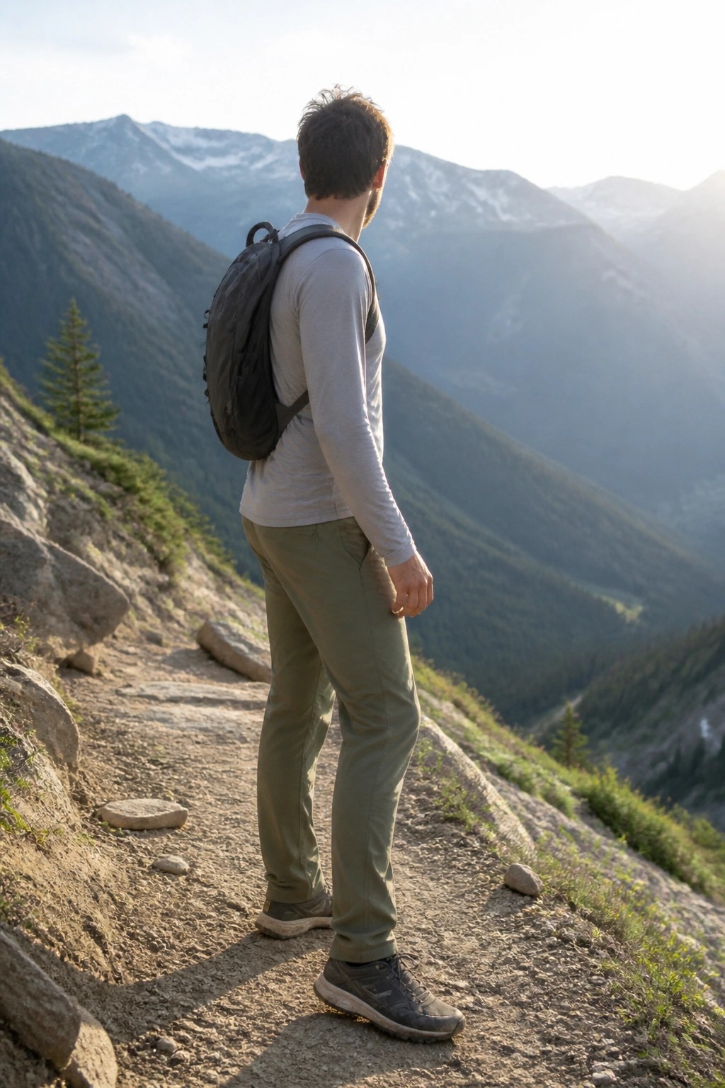 Man in olive green rugged travel pants, light gray long sleeve shirt, black backpack, and dark hiking shoes standing on rocky trail.