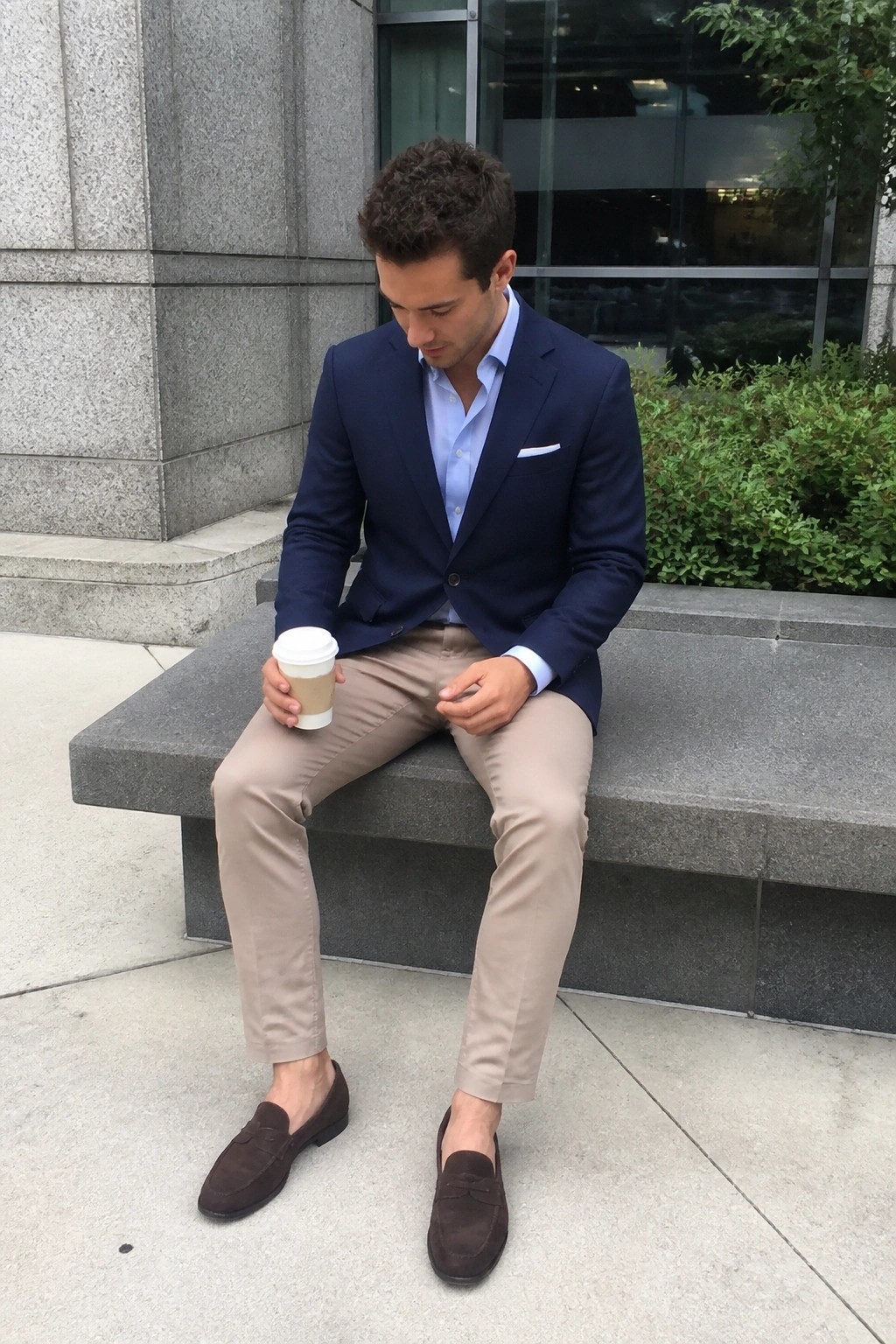 Man in navy blazer and beige pants holding coffee on stone bench