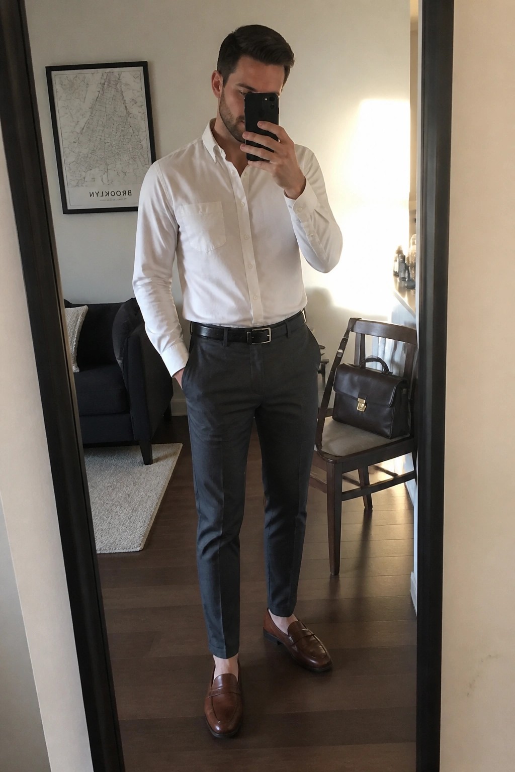 Man in white shirt and gray pants taking mirror selfie with phone.