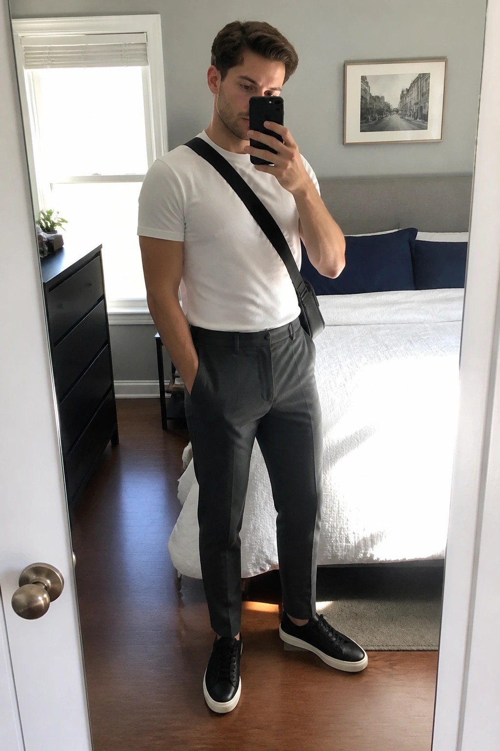 Man in white t-shirt and gray pants taking mirror selfie in bedroom
