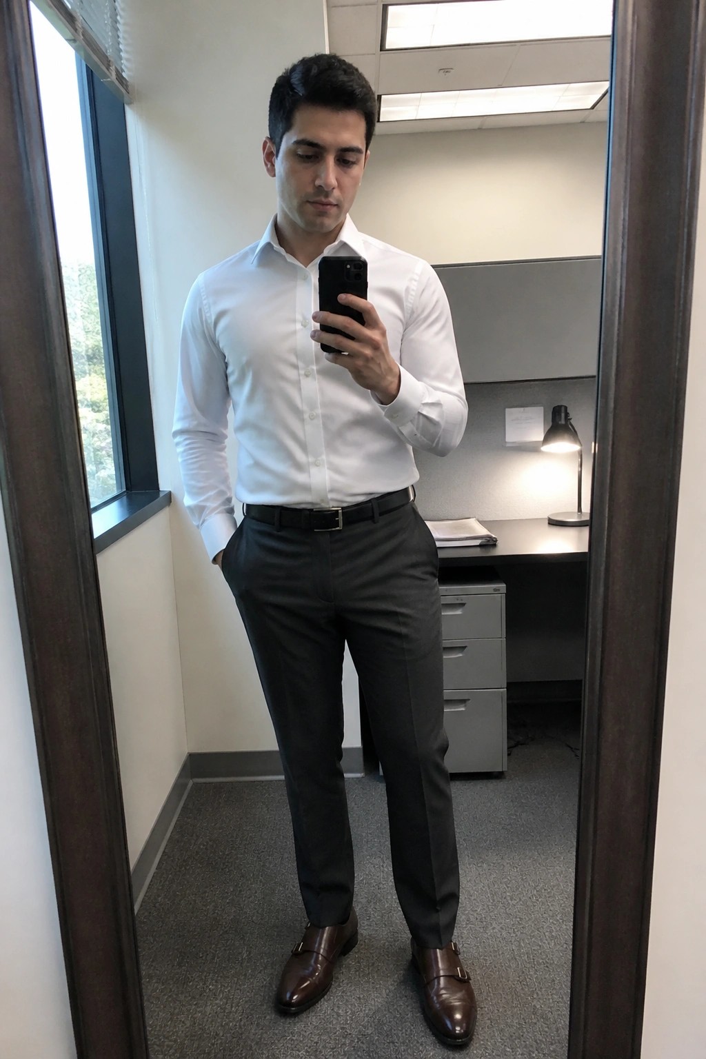 Man in white shirt and gray pants taking mirror selfie in office