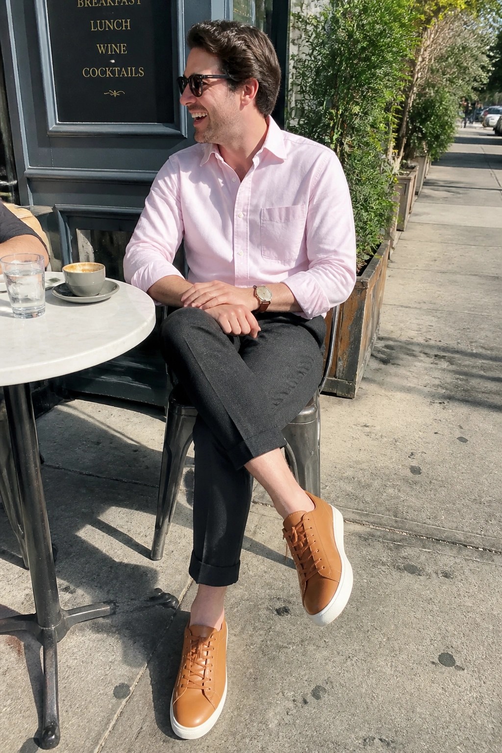Man in pink shirt and brown sneakers smiles at outdoor cafe table