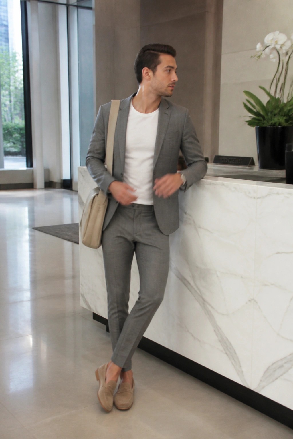 Man wearing gray suit pants and blazer over a white t-shirt with beige loafers and a crossbody bag standing by a marble counter.