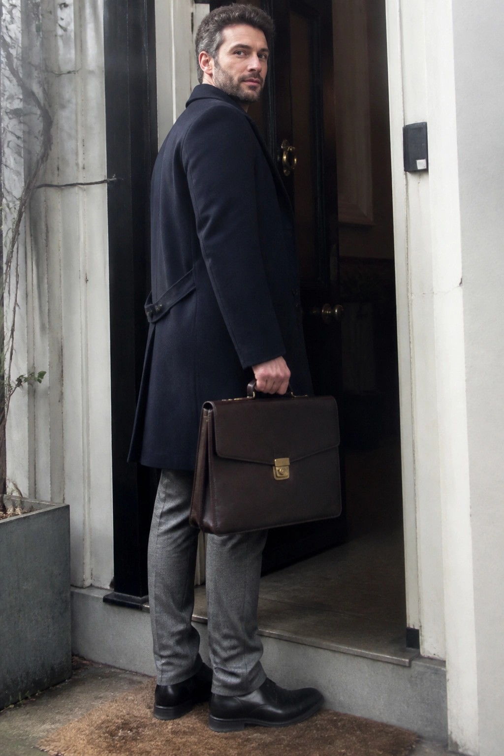 Man in navy coat holding brown briefcase stands in doorway looking back