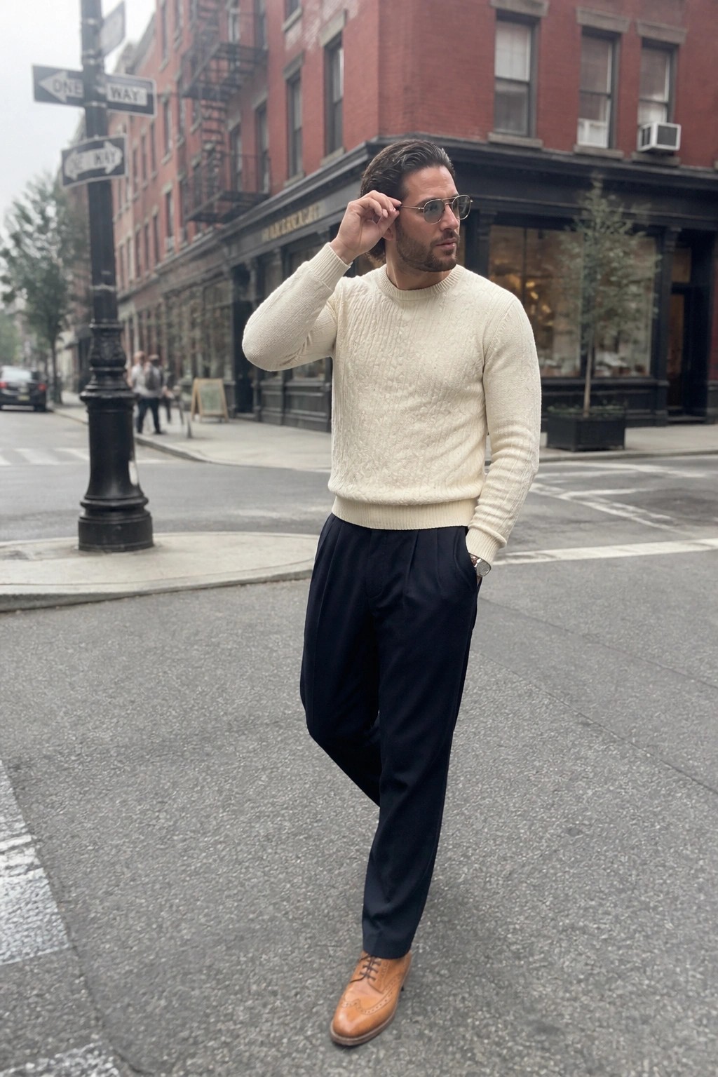 Man in cream cable-knit sweater and navy trousers adjusting sunglasses on city street