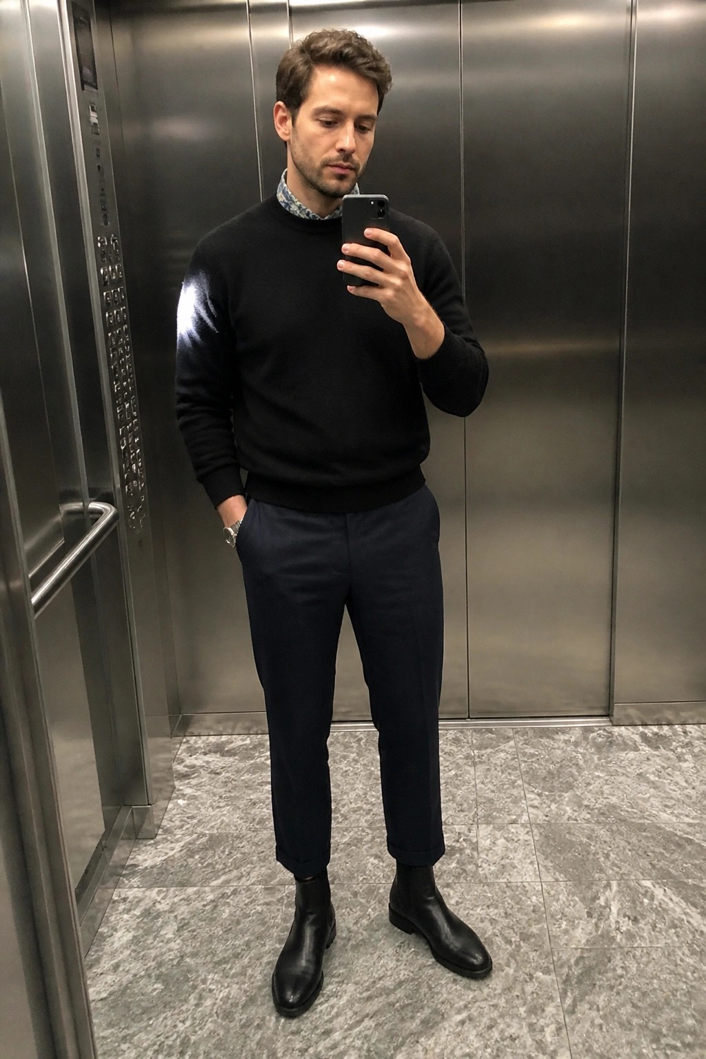 Man in black sweater taking mirror selfie inside metallic elevator with navy pants and boots.