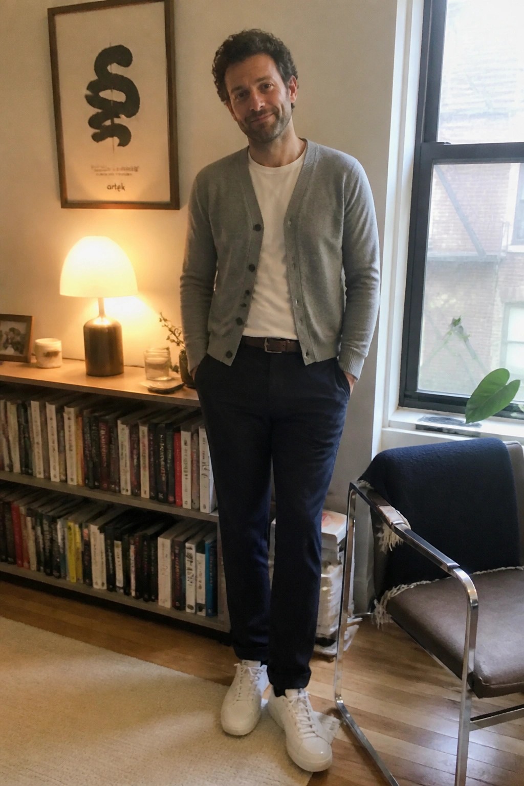 Man wearing navy dress pants, gray cardigan over white t-shirt, brown belt, and white sneakers in an indoor setting with bookshelves and a window.