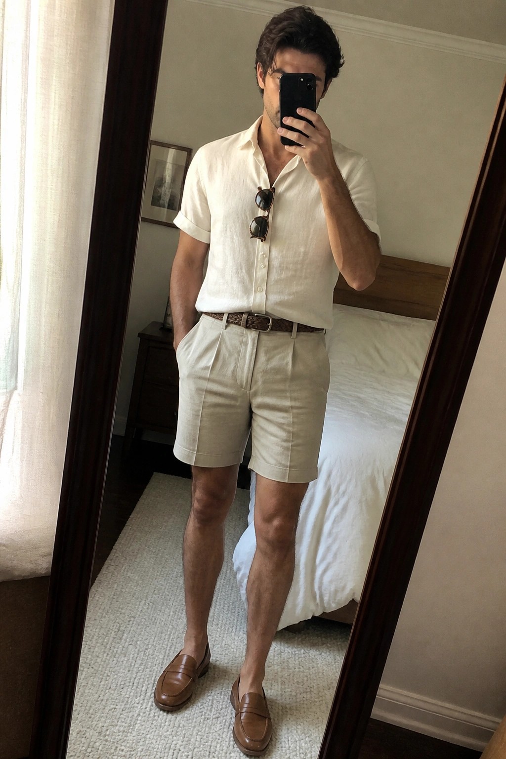 Man wearing cream short-sleeve linen button-up shirt tucked into pleated beige shorts with brown woven belt and brown leather loafers while holding a phone in a bedroom mirror selfie.
