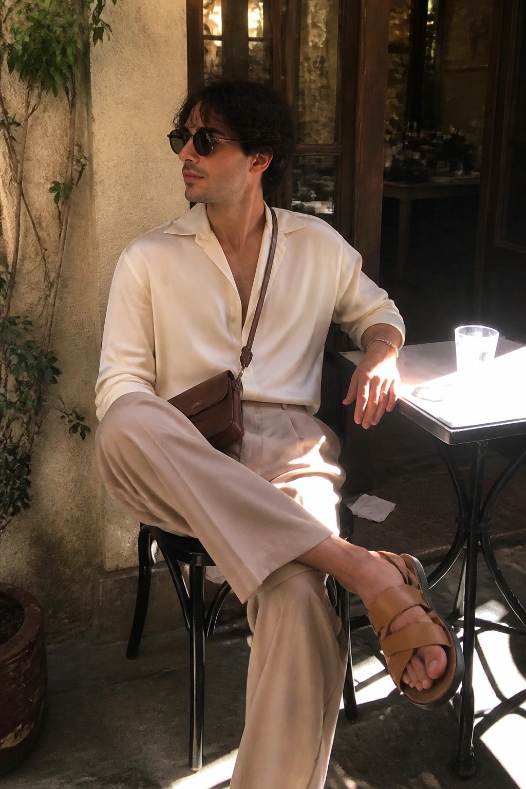 Man seated outdoors wearing an off-white button-down shirt, light beige wide-leg trousers, brown leather double-strap sandals, and a brown leather crossbody bag.