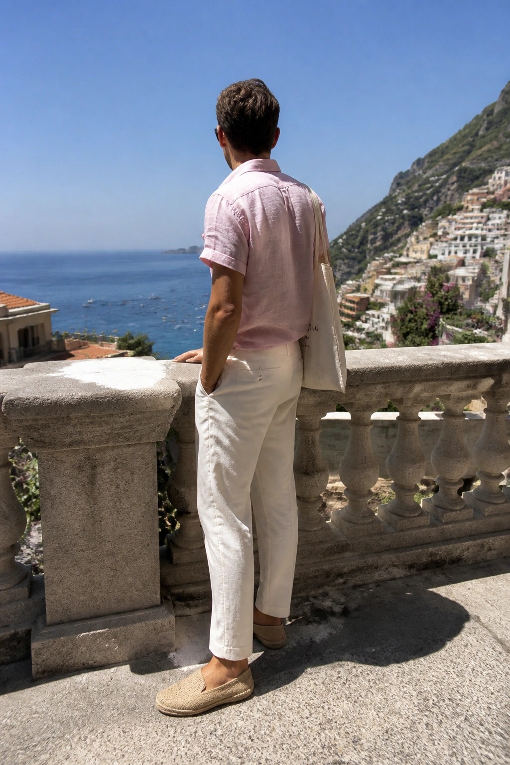 Man in pink shirt and white pants overlooking blue sea and hillside village.
