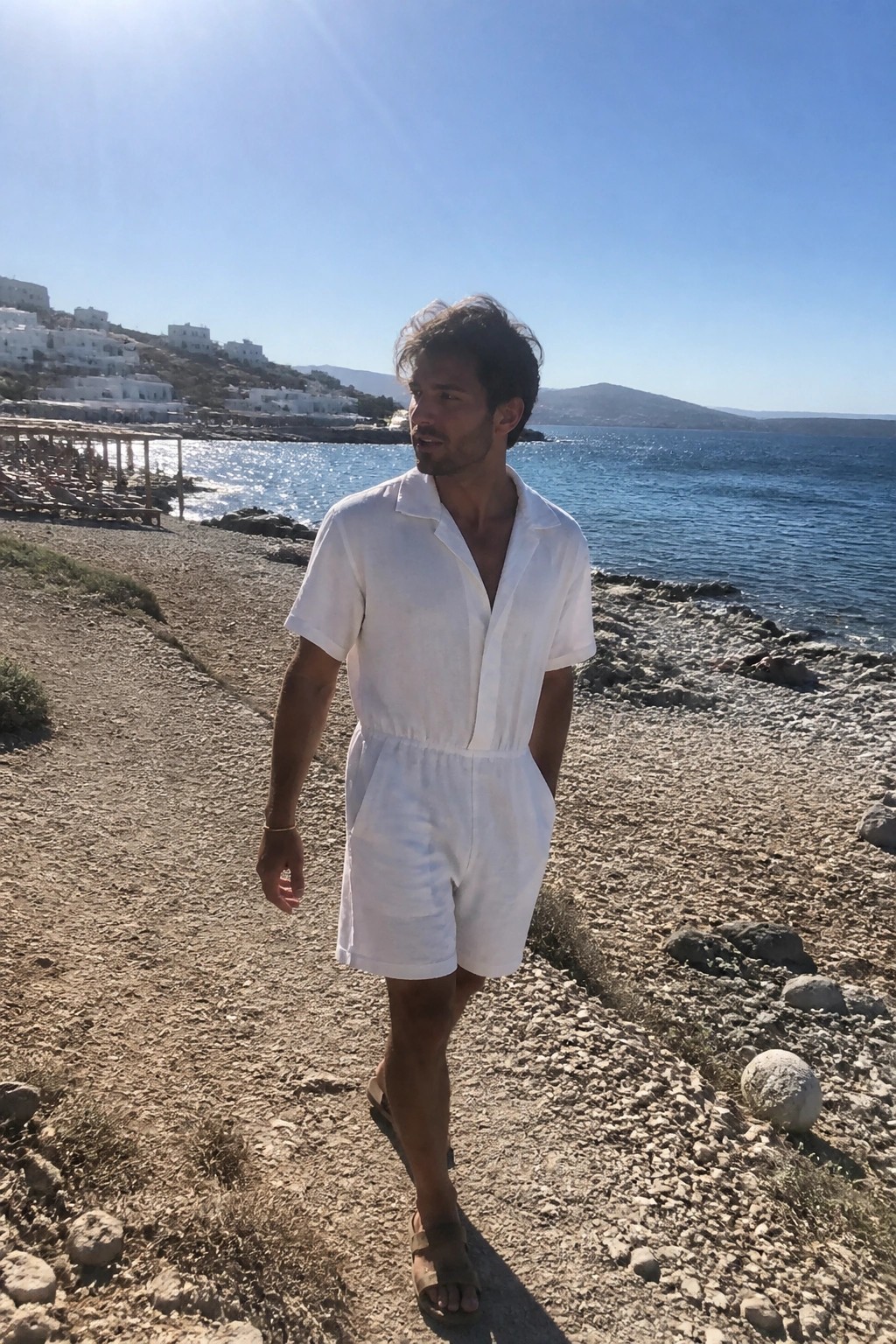 Man in white short-sleeved romper walking on rocky beach by blue sea