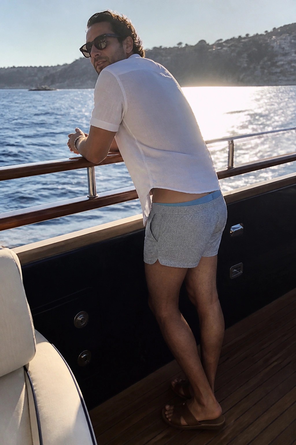 Man in white shirt and patterned shorts leaning on yacht railing over water
